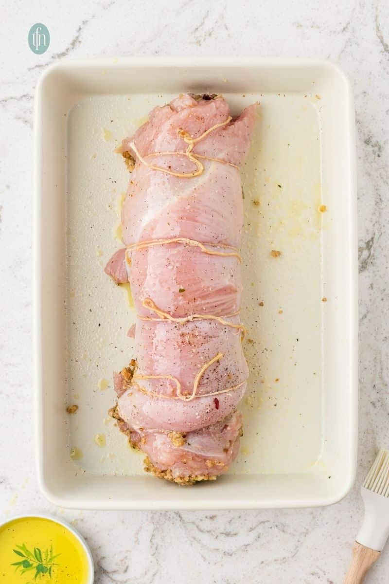 An overhead shot of the raw stuffed turkey breast, rolled up and tied with kitchen twine, resting in a white rectangular baking dish, lightly coated with oil.