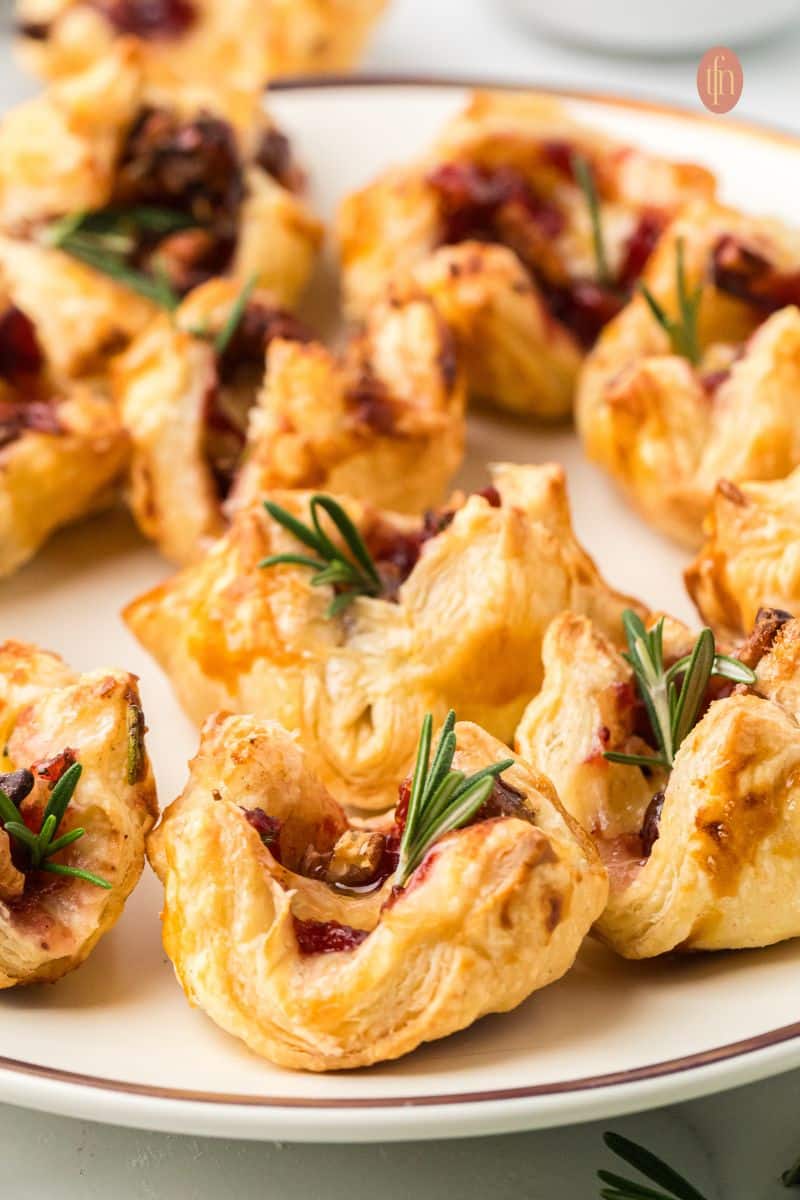 Low-angle, close-up view of several Brie and cranberry appetizers arranged on a white plate with a brown rim, showing the flakiness of the pastry and the melted cheese filling.