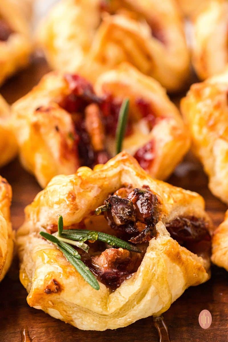 Tight close-up of a single golden brown Brie and cranberry puff pastry bite, garnished with a fresh sprig of rosemary and drizzled with honey.