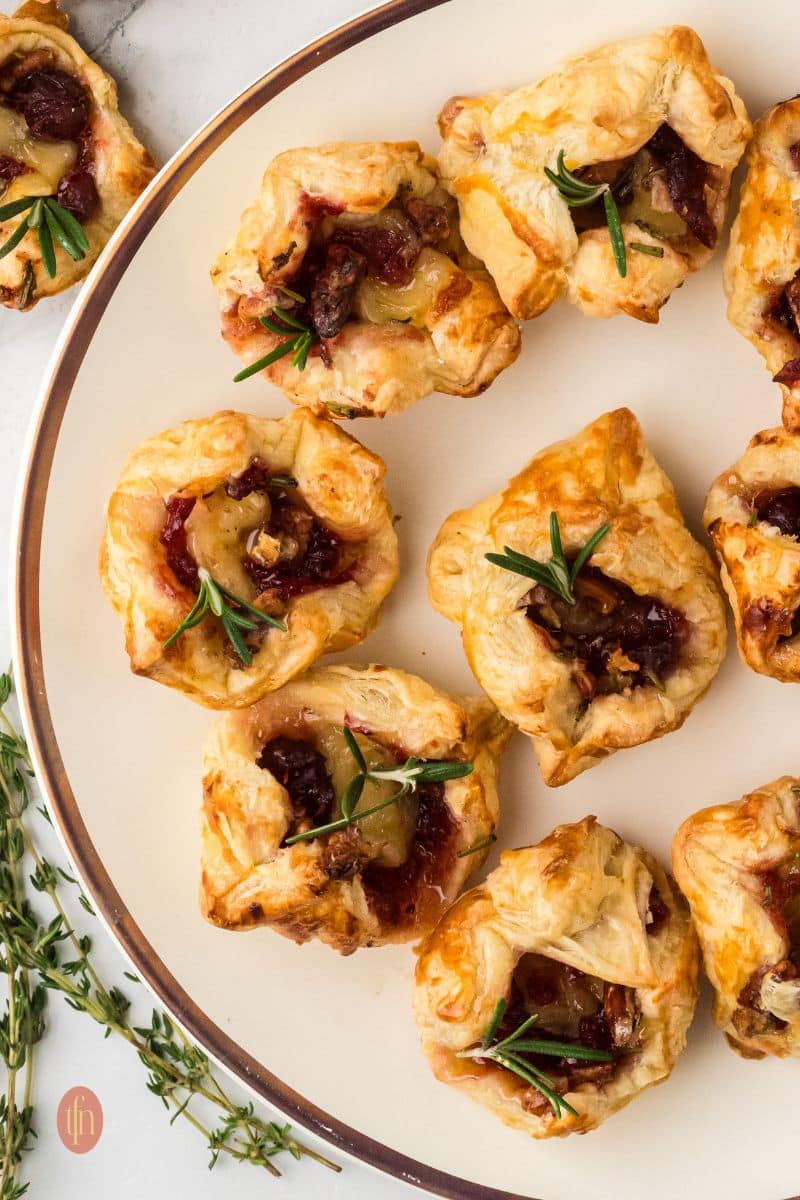 Extreme close-up shot of multiple baked Brie bites on a wooden board, showcasing the melted Brie cheese, red cranberry sauce, and rosemary sprigs nested in the flaky puff pastry cups.