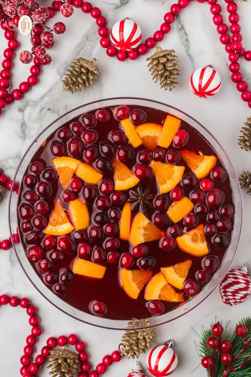 Overhead view of a large glass bowl of holiday punch garnished with floating cranberries, orange slices, and a star of anise. The bowl is surrounded by Christmas decor like gold pinecones and red bead garlands.