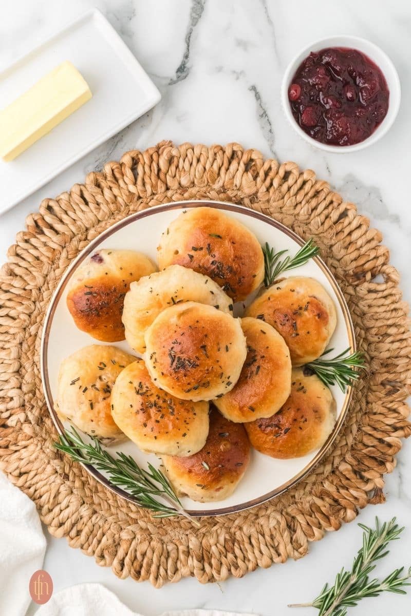 a plate of easy dinner rolls stacked with rosemary sprigs for garnish with a bowl of cranberries and butter for serving.