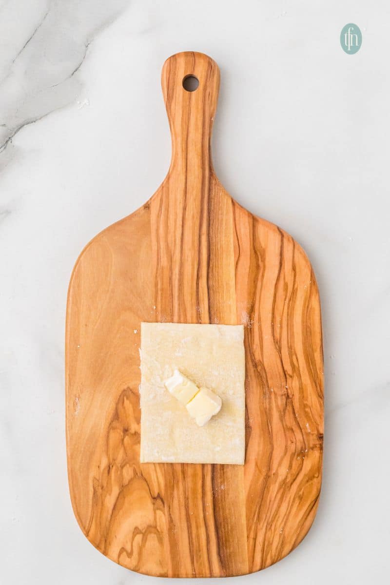 Overhead preparation shot on an olive wood cutting board: a square of puff pastry dough topped with two small cubes of Brie cheese.