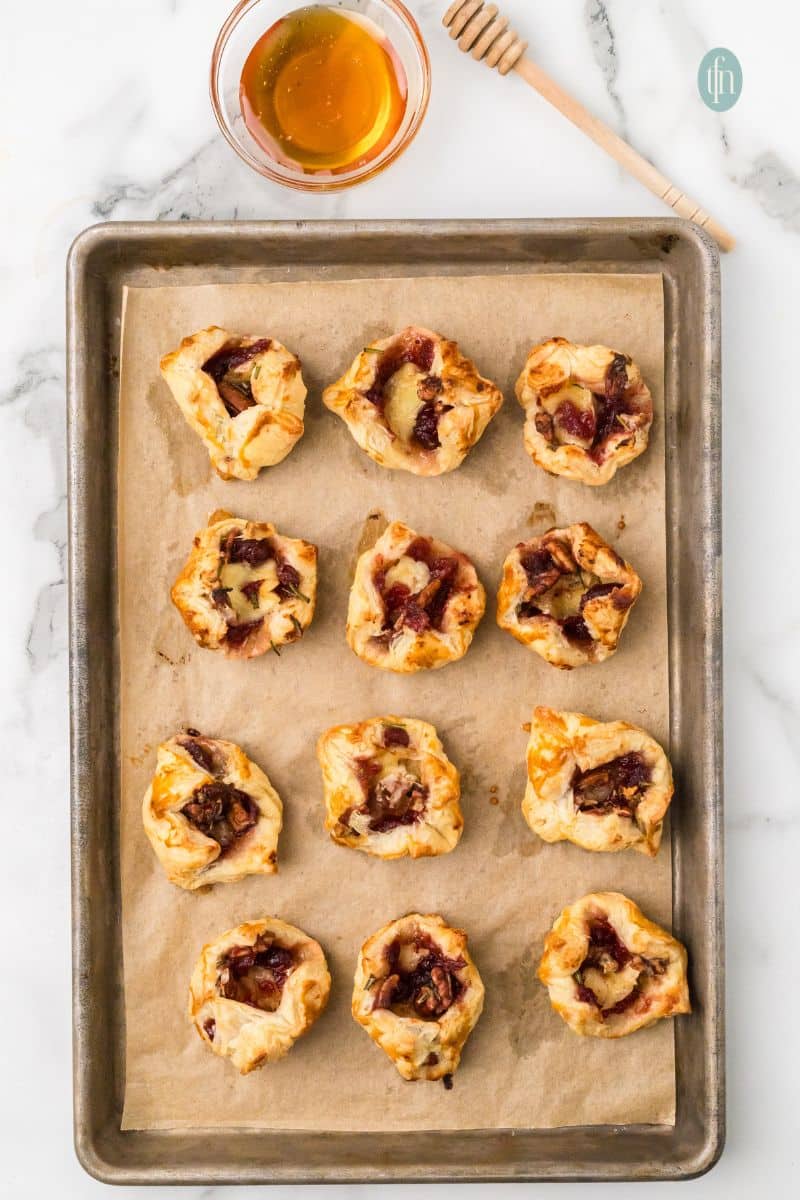 Overhead view of a baking sheet with twelve baked Brie and cranberry bites. A small bowl of honey and a wooden dipper are visible at the top, ready for drizzling.