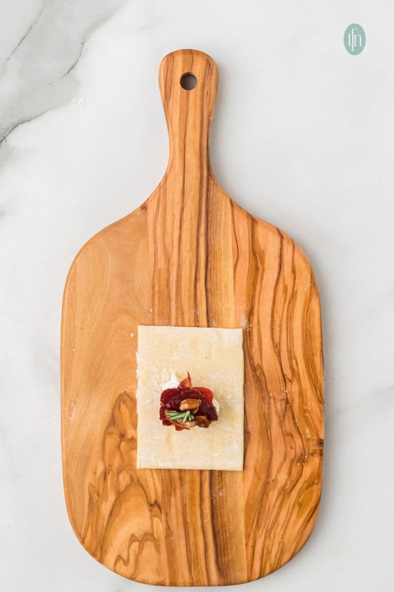 Overhead preparation shot on a wooden cutting board: a square of puff pastry is fully assembled with Brie cheese, cranberry sauce, pecans, and a small sprig of rosemary.
