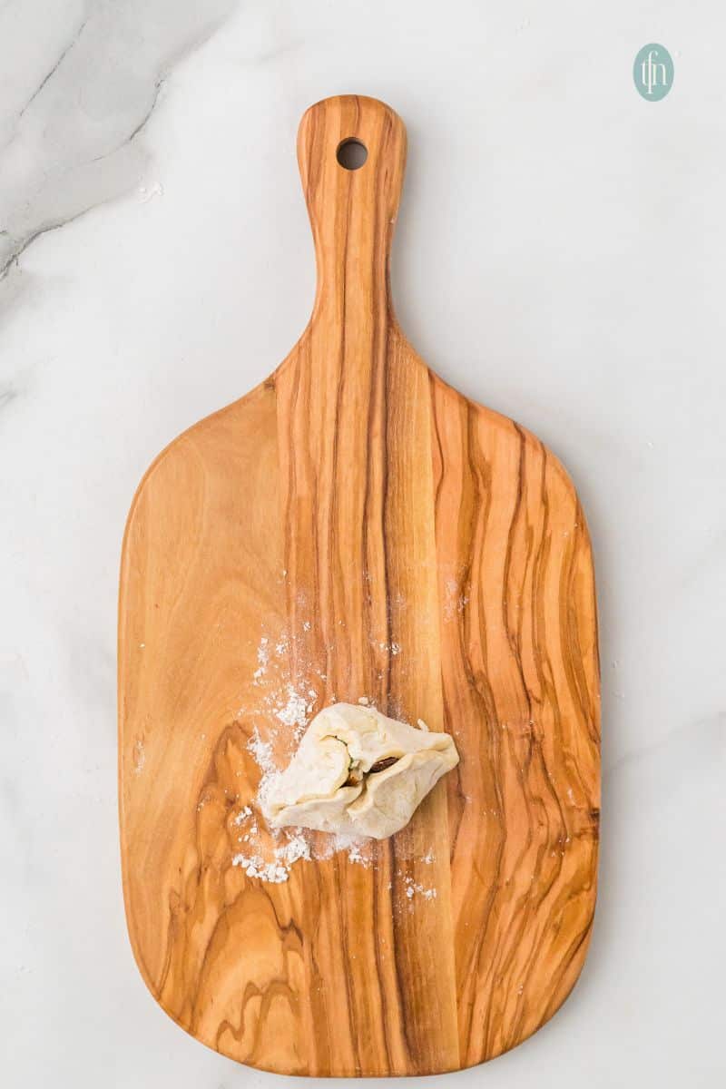 Overhead preparation shot on a wooden cutting board, showing a single Brie bite carefully pinched and folded into a small, sealed pouch shape.