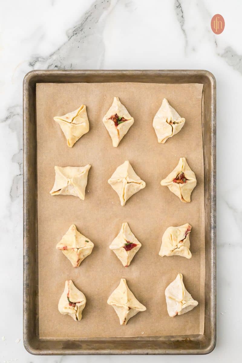 Overhead view of a baking sheet lined with parchment paper, holding twelve uncooked, filled Brie bites folded into pouch shapes, ready for the oven.