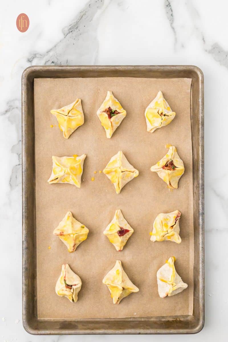 Overhead view of the baking sheet with twelve uncooked Brie bites that have been lightly brushed with an egg wash for a golden brown finish.