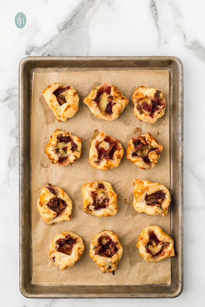 Overhead view of a baking sheet with twelve golden brown Brie and cranberry bites that are fully baked and puffed up, with the melted cheese and cranberry filling visible.