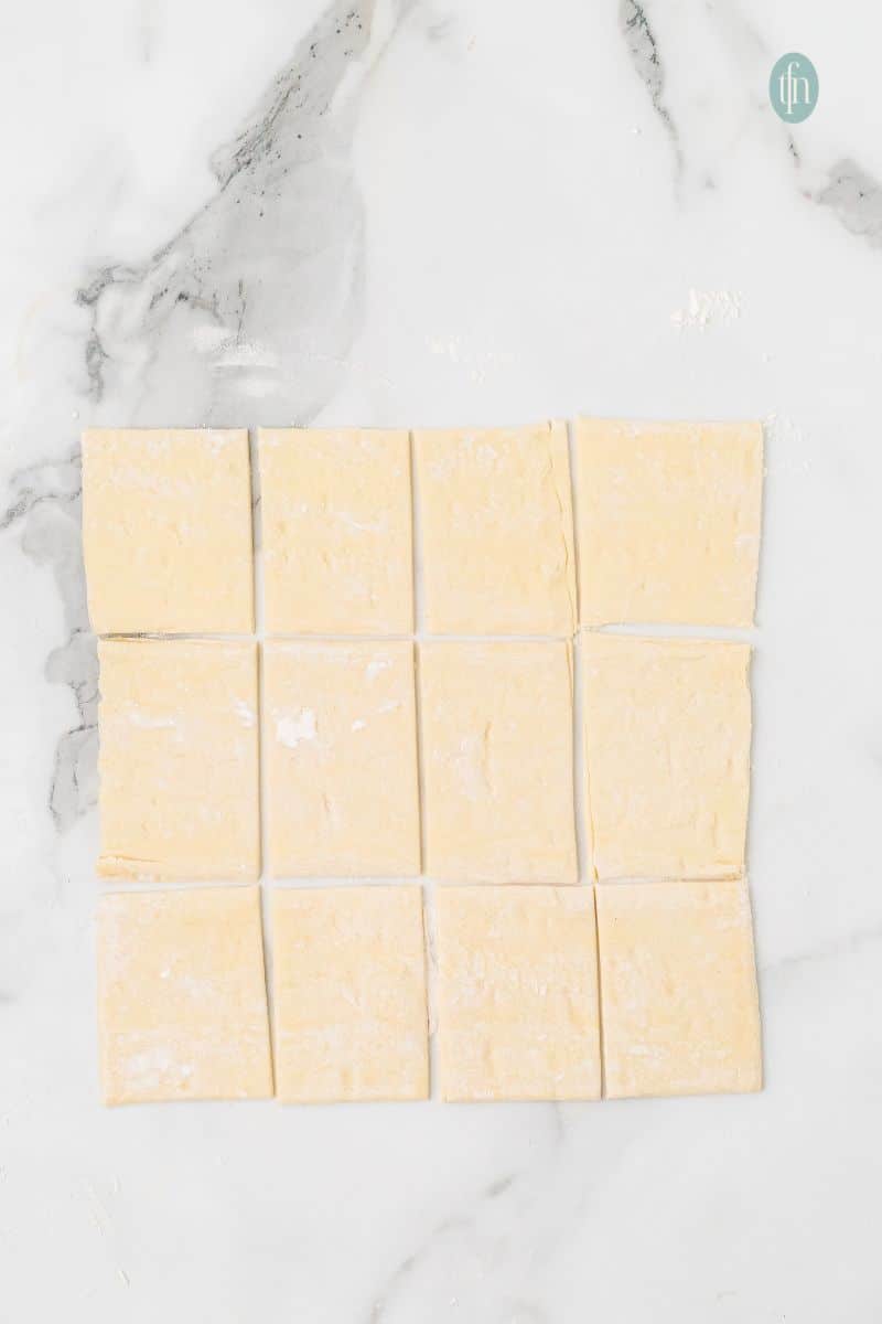 Overhead shot of a sheet of raw puff pastry dough, cut into twelve uniform squares, resting on a marble countertop.