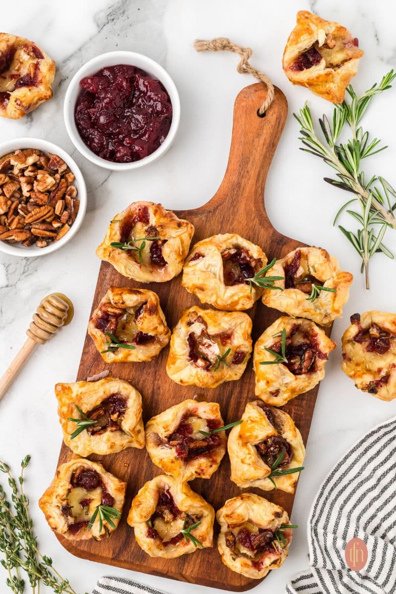 Top-down flat lay of the baked Brie and cranberry appetizers served on a rustic wooden board, accompanied by bowls of cranberry sauce and chopped pecans, and a honey dipper.