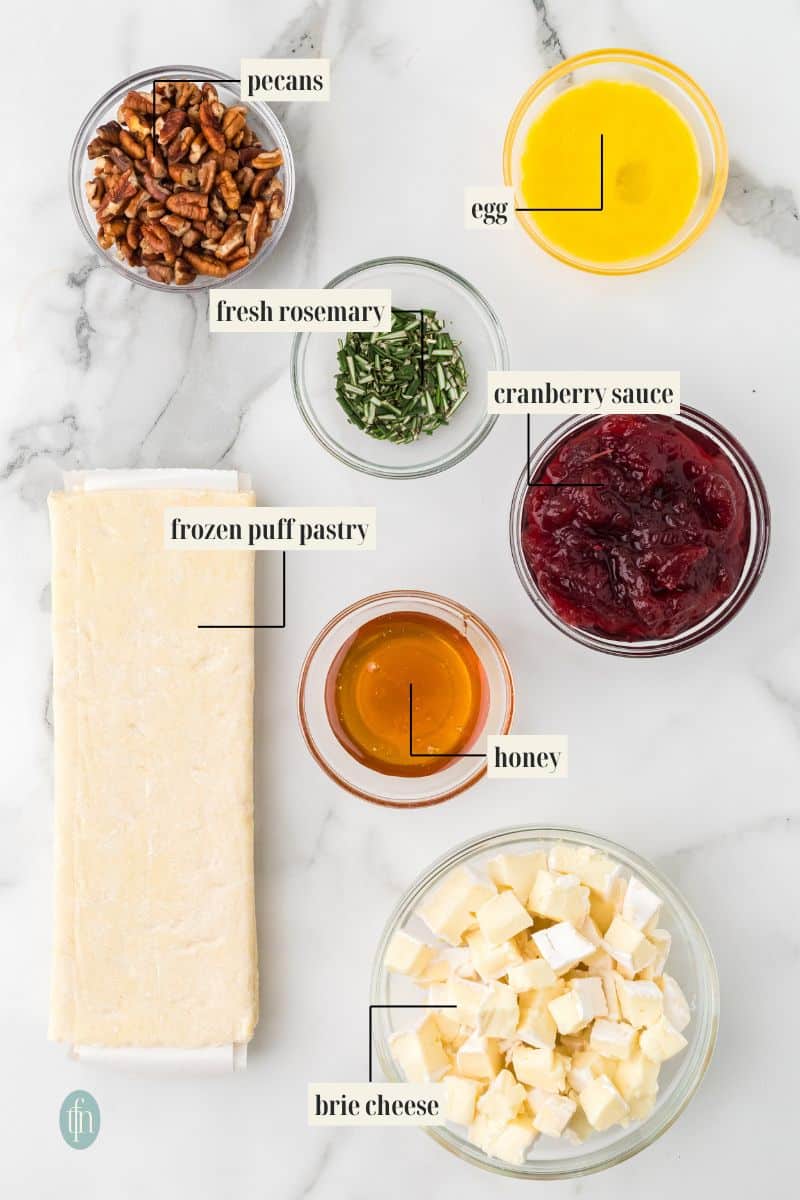 Flat lay of all the raw ingredients for the appetizer, with labels: frozen puff pastry, chopped brie cheese, cranberry sauce, pecans, chopped fresh rosemary, a beaten egg, and honey.