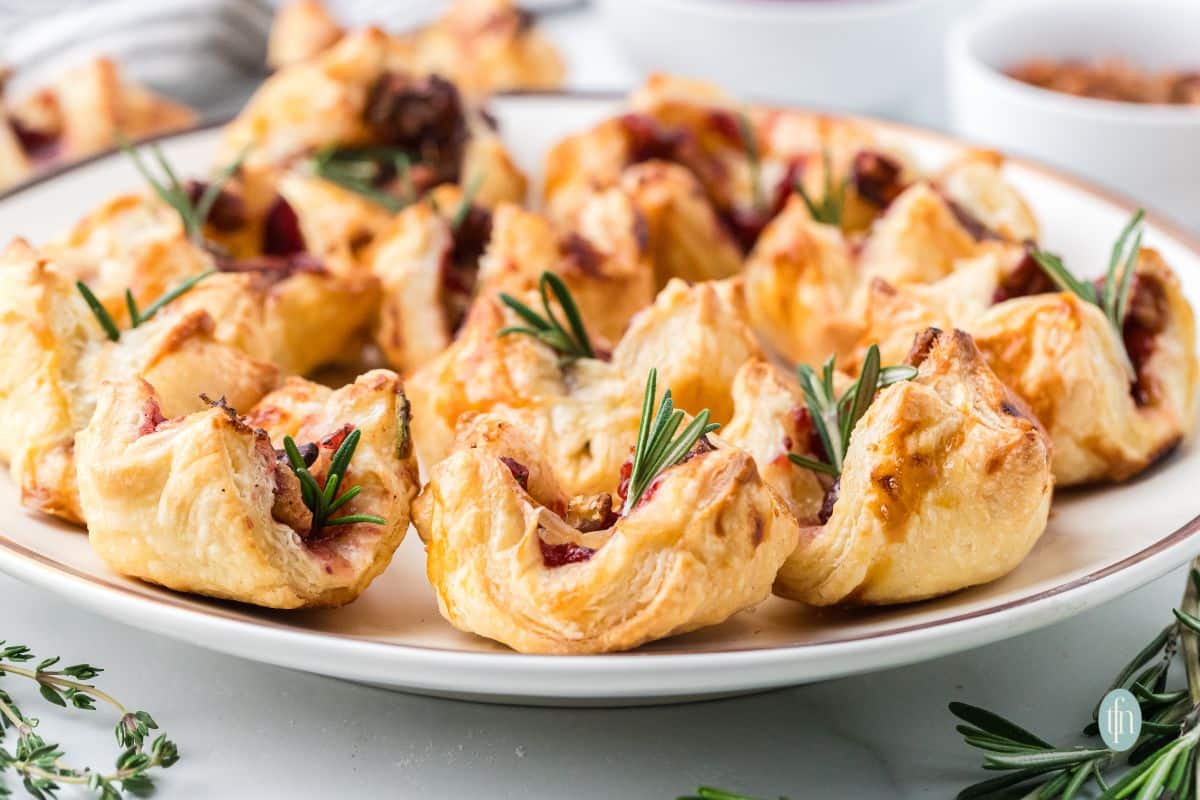 Close-up, horizontal view of a platter piled high with flaky, golden Cranberry Brie Bites, each topped with a small sprig of rosemary.