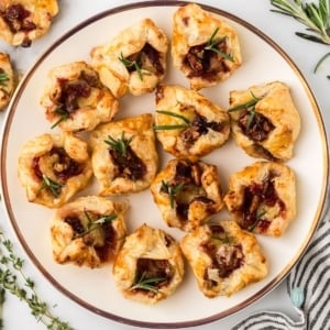 Overhead square photo of twelve golden brown, individual Cranberry Brie Bites topped with fresh rosemary, served on a round white plate with a gold rim.