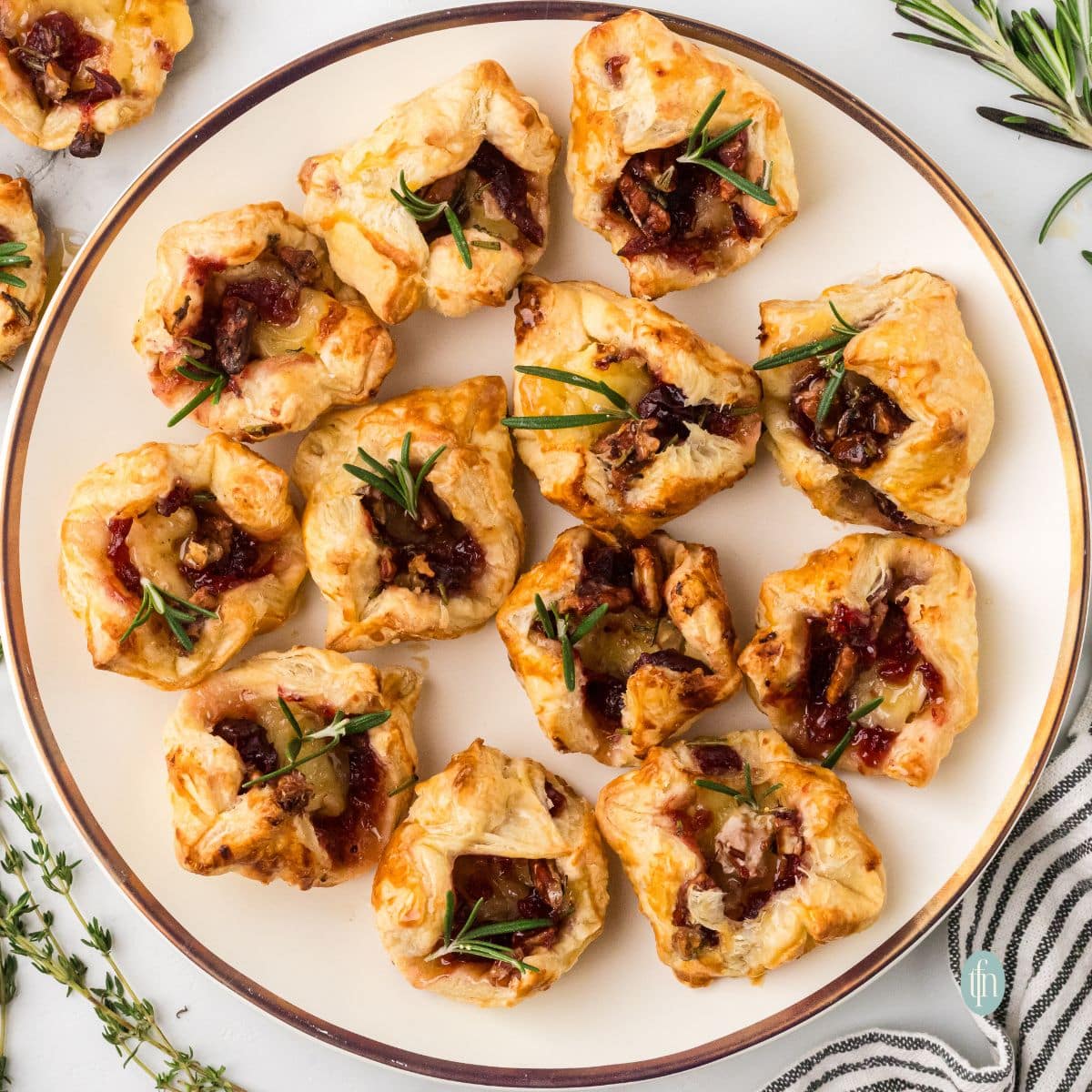 Overhead square photo of twelve golden brown, individual Cranberry Brie Bites topped with fresh rosemary, served on a round white plate with a gold rim.