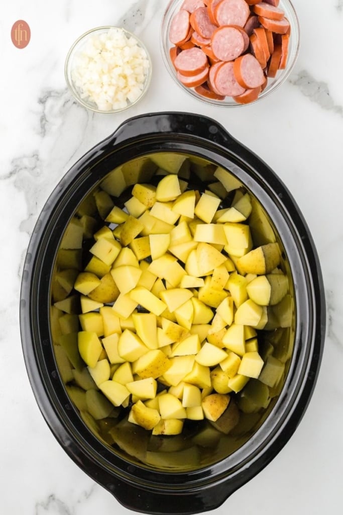 a prep image of a crockpot full of cubed potatoes with a bowl of sausage and chopped onions next to it.