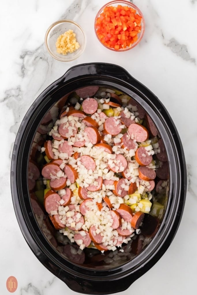 a prep image of a crockpot full of cubed potatoes, chopped onion, and sausage with a bowl of red bell pepper and garlic in bowls next to it.
