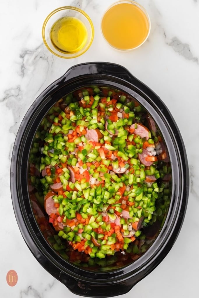 a prep image of a crockpot full of cubed potatoes, sausage, onion, red bell pepper, green bell pepper, and garlic with bowls of broth and oil next to it.