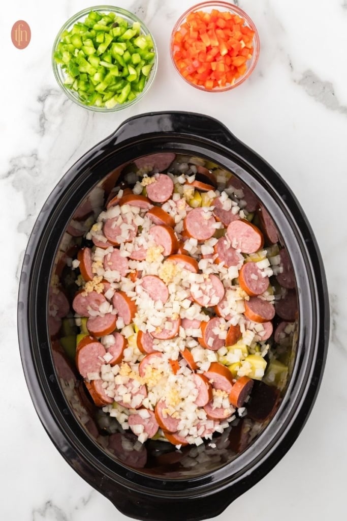 a prep image of a crockpot full of cubed potatoes, sausage, onion, red bell pepper, green bell pepper, and garlic.