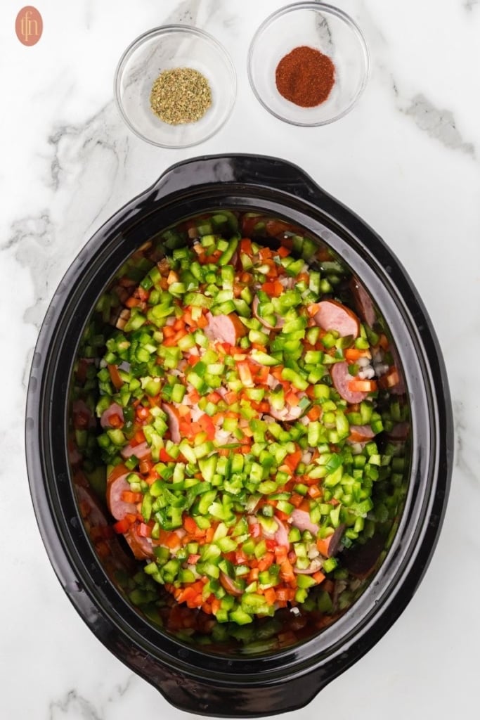 a prep image of a crockpot full of cubed potatoes, sausage, onion, red bell pepper, green bell pepper, and garlic with bowls of spices above it.