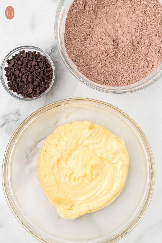 a bowl of dough mixture with a bowl of cocoa powder and mini chocolate chips on the side.