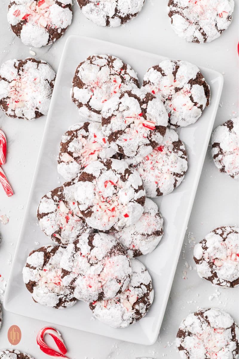 a plate of chocolate crinkle cookies with more cookies and candy cane on the side.