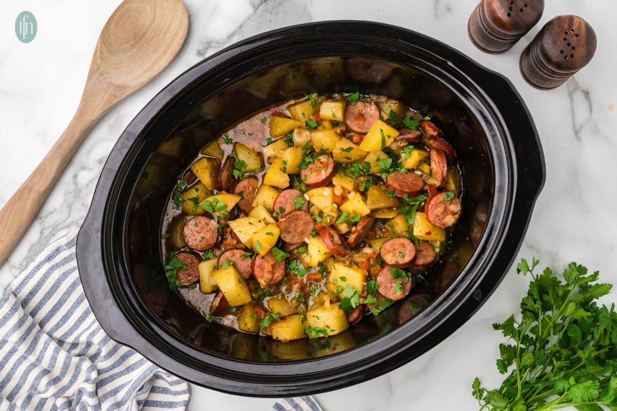 a horizontal image of sausages and potatoes in slow cooker in a crock pot with salt and pepper shakers, a wooden spoon and parsley next to it.