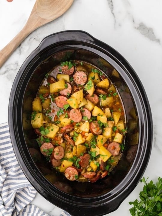 a prep image of a crockpot full of cubed potatoes, sausage, onion, red bell pepper, green bell pepper, garlic, and spices with a wooden spoon next to the crock pot and parsley.