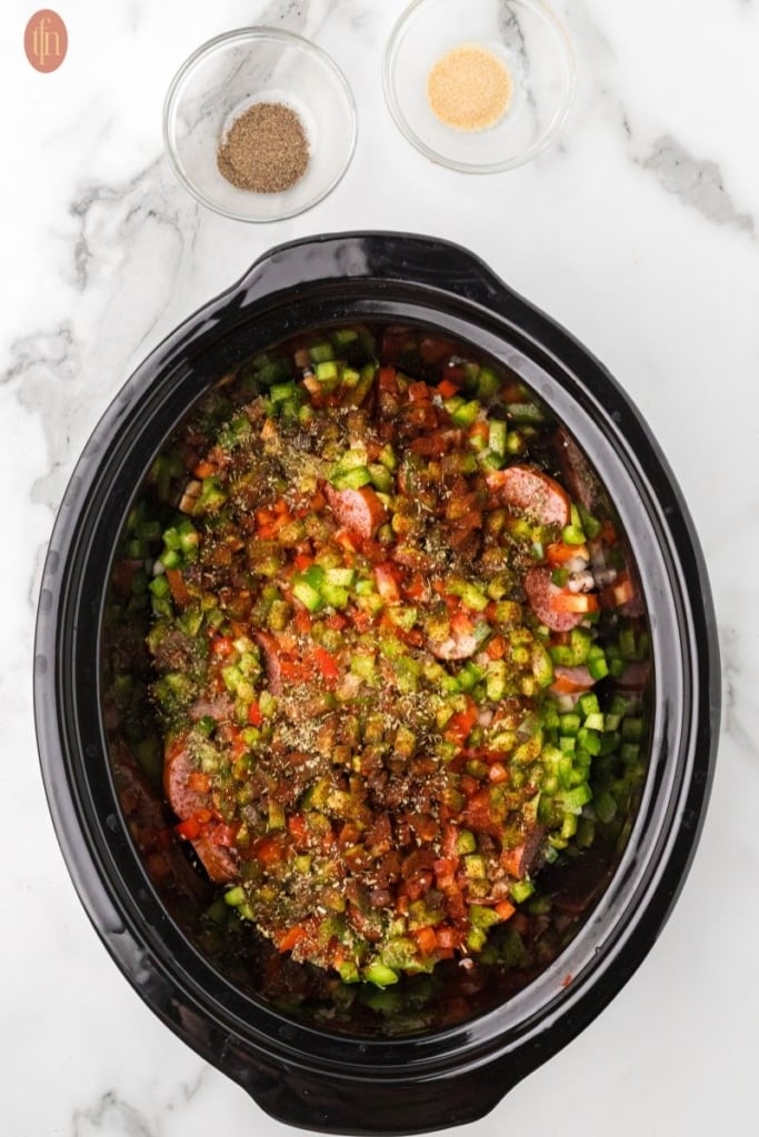 a prep image of a crockpot full of cubed potatoes, sausage, onion, red bell pepper, green bell pepper, and garlic with bowls of spices above it.