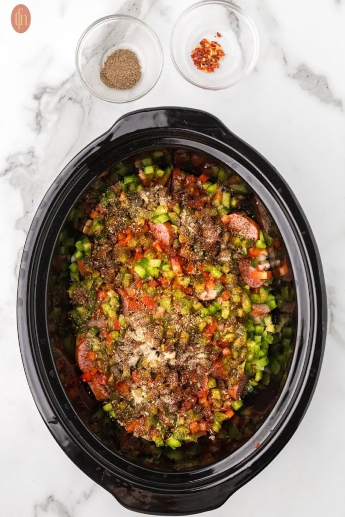 a prep image of a crockpot full of cubed potatoes, sausage, onion, red bell pepper, green bell pepper, and garlic with bowls of spices above it.