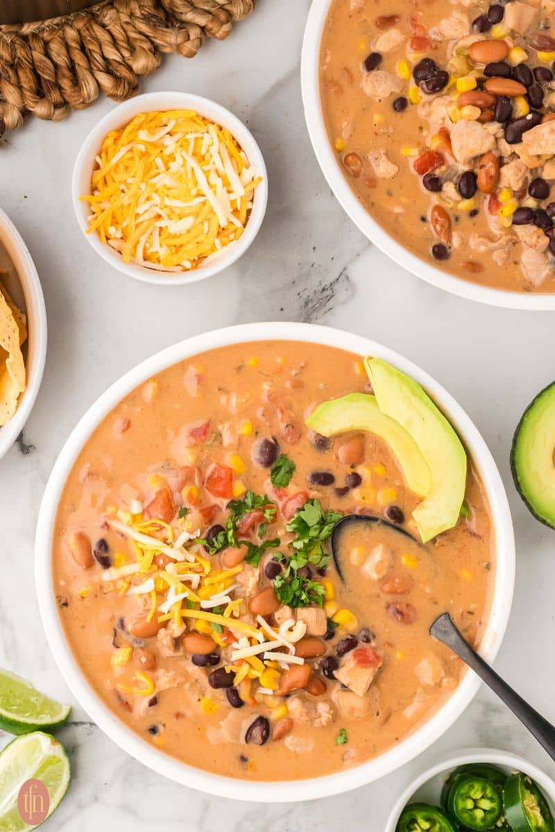 two white bowls of chicken taco soup with tortilla chips, lime, jalapenos, avocado and extra cheese on the side.