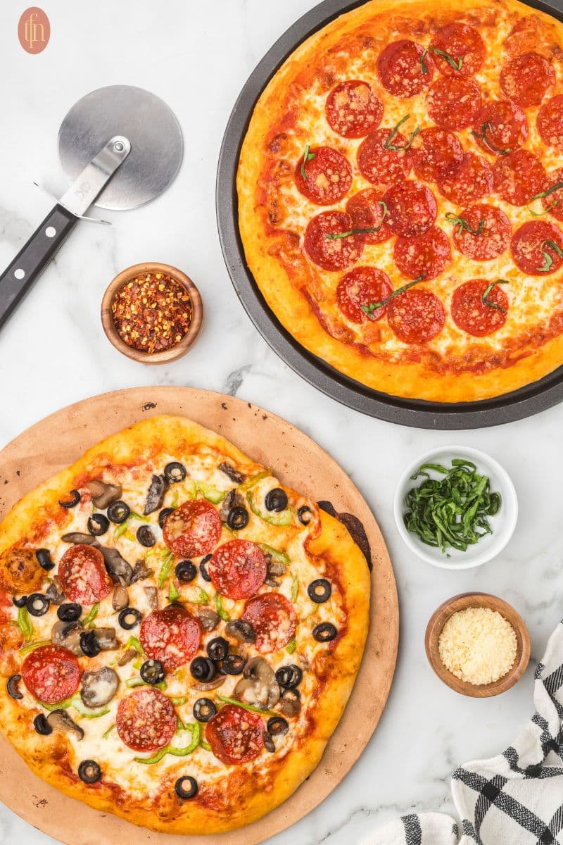 a two different homemade pizza, on the lower left side, it's a pizza topped with pepperoni and black olives while the other pizza on the upper right side topped with pepperoni. It also has a three small seasoning bowls and a pizza cutter on the side.