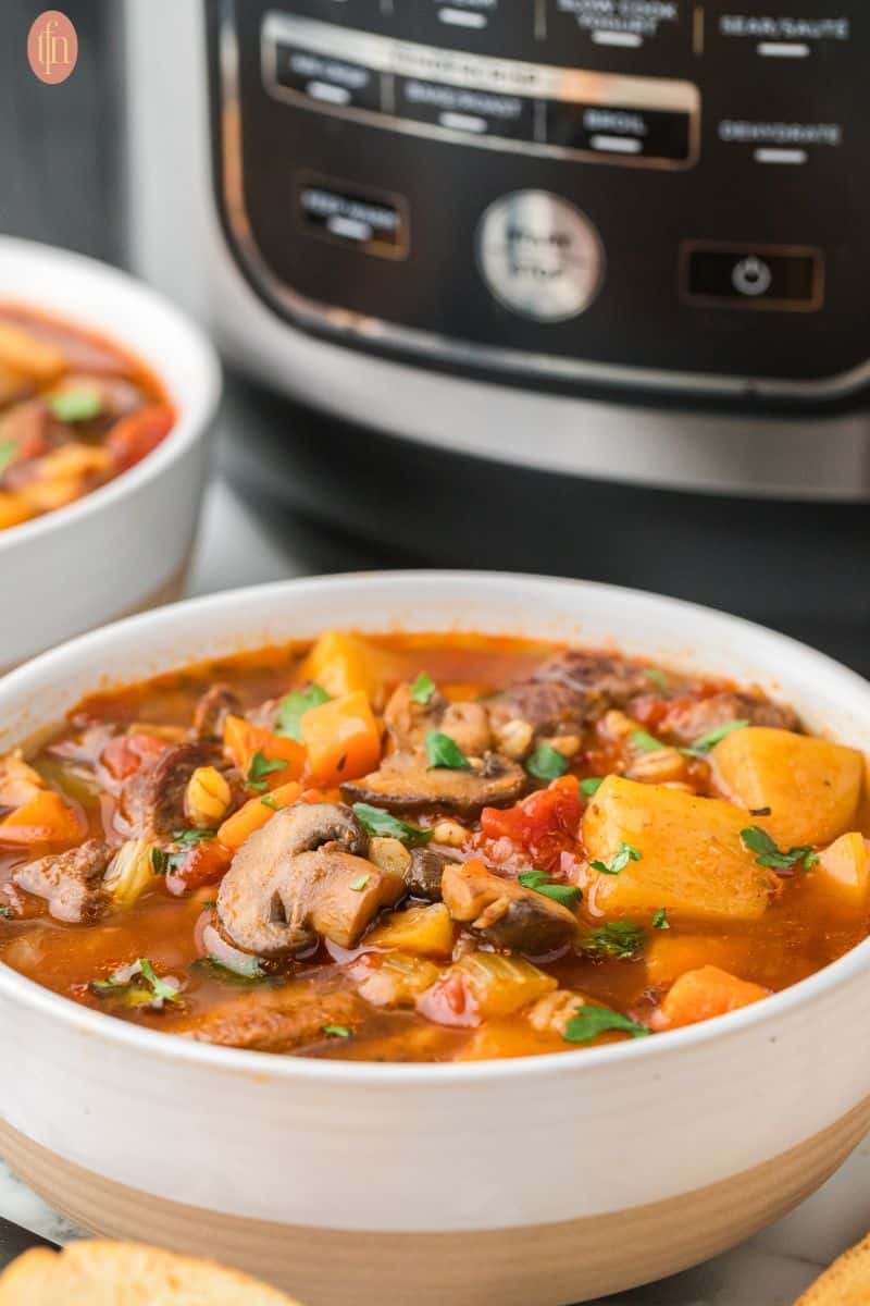 a close up shot of instant pot beef and barley soup in a bowl and an instant pot at the back.
