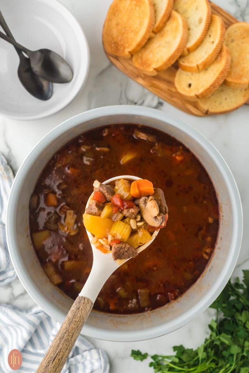 a close-up shot of beef and barley soup in a scoop and more soup in a pot in the background. It also has a bread and a small bowl with two spoons in it.