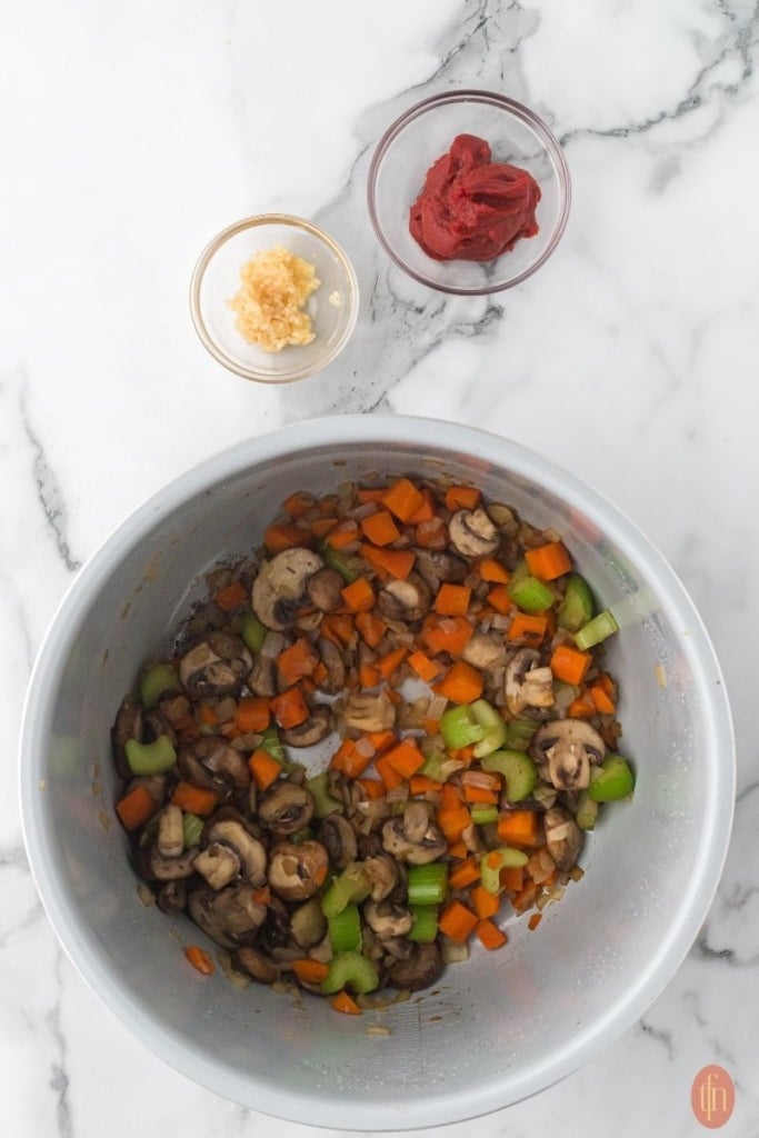 a pot of saut&eacute;ed carrots, celery, mushrooms, and onion with two small bowls of garlic and tomato paste on the side.