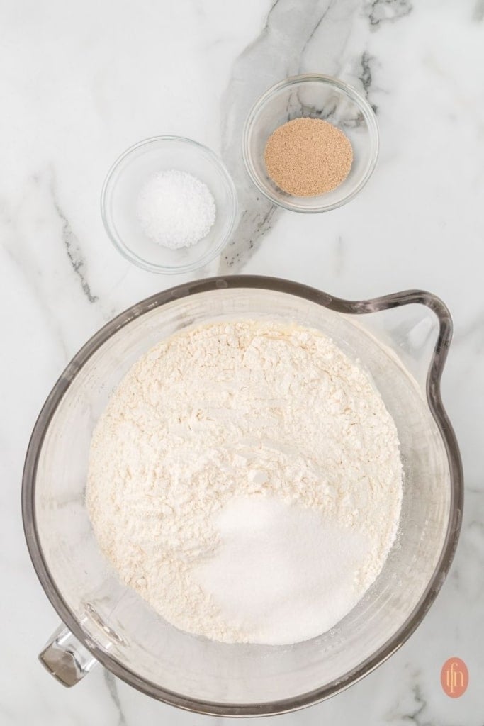 a dough on a stand mixer with a bowl of yeast and a granulated sugar on the side.
