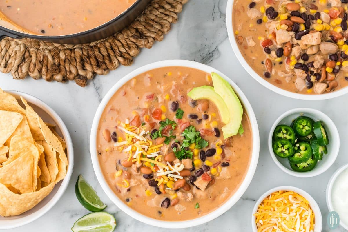 overhead view of a chicken taco soup in white bowl with tortilla chips, lime, jalapenos and extra cheese on the side.