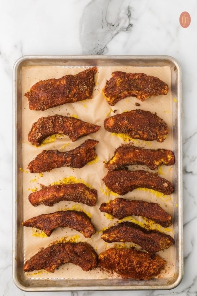 slices of pork ribs coated with yellow mustard and rib seasoning on a sheet pan.