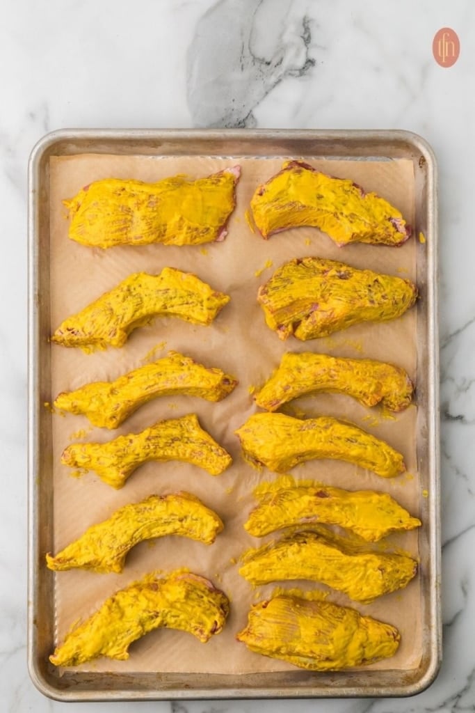 slices of pork ribs coated with yellow mustard on a sheet pan.