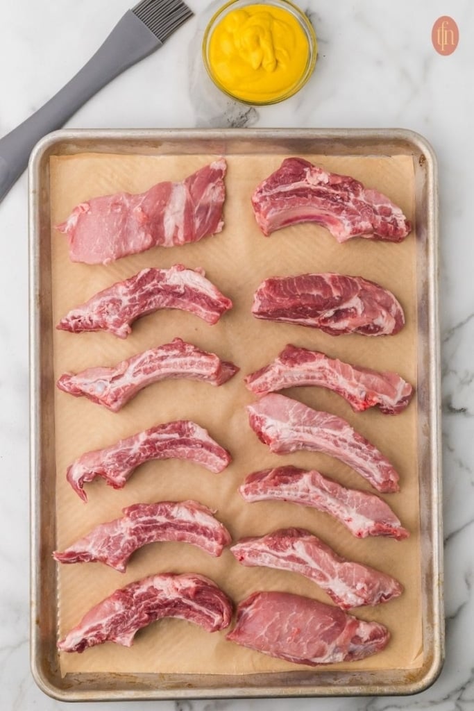 a raw pork ribs on a sheet of parchment paper with a bowl of yellow mustard and brush on the side.