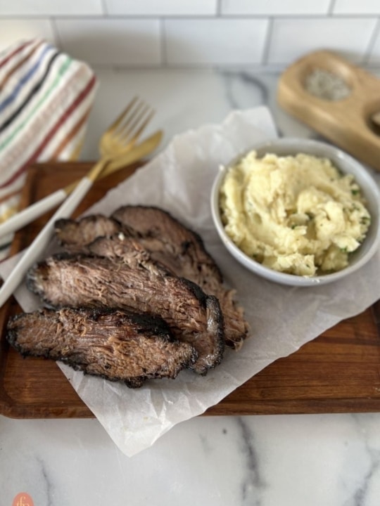 a bowl of mashed potatoes served alongside sliced smoked brisket on a wooden cutting board.