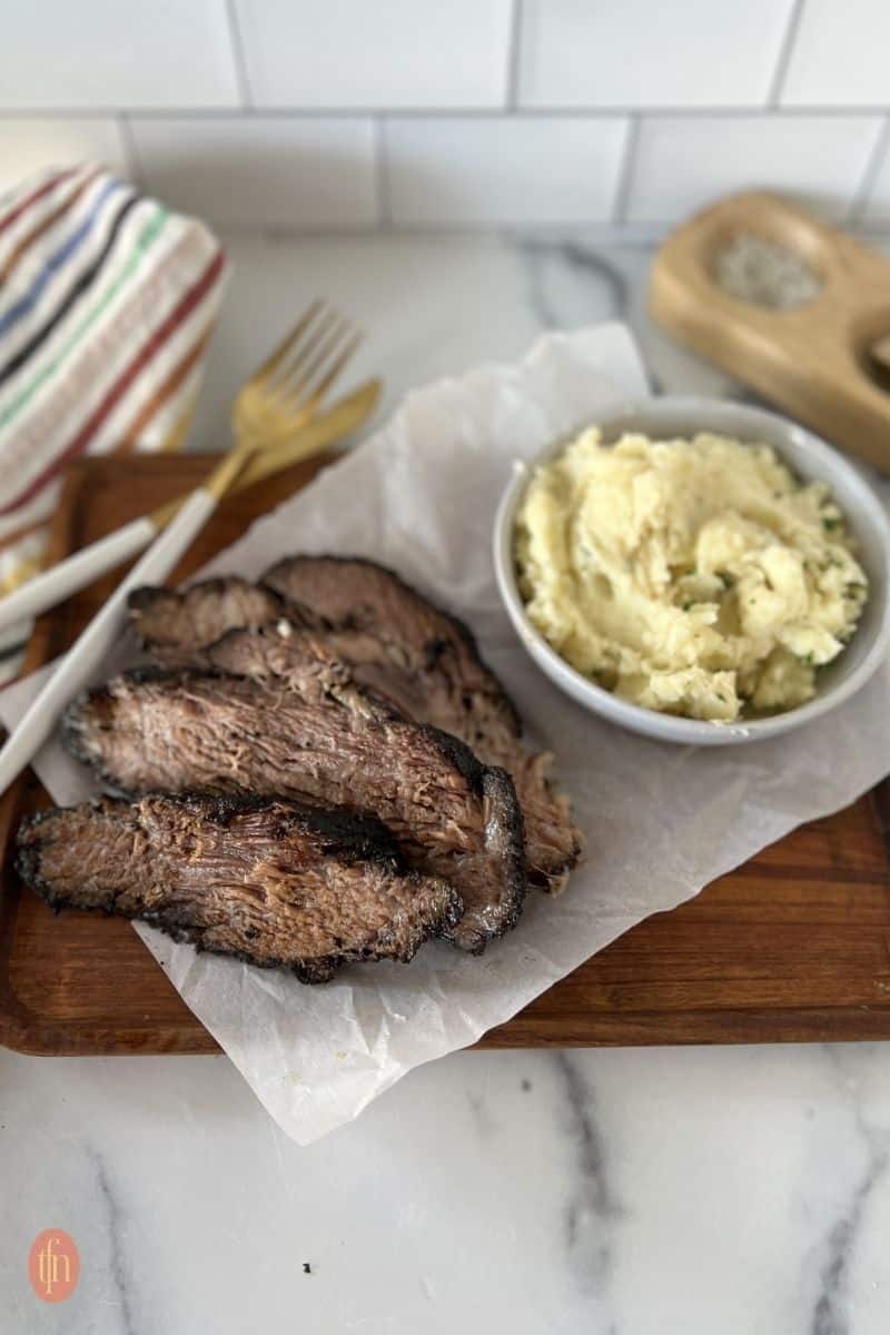 a bowl of mashed potatoes served alongside sliced smoked brisket on a wooden cutting board.