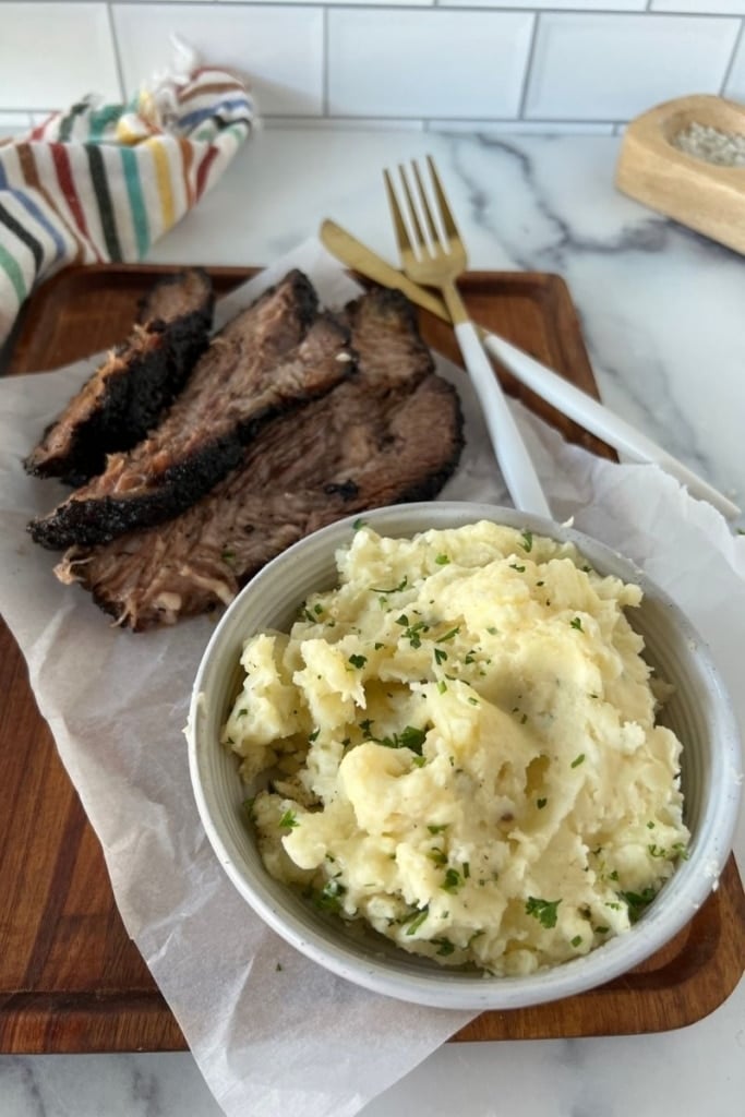 a bowl of mashed potatoes served alongside sliced smoked brisket on a wooden cutting board.