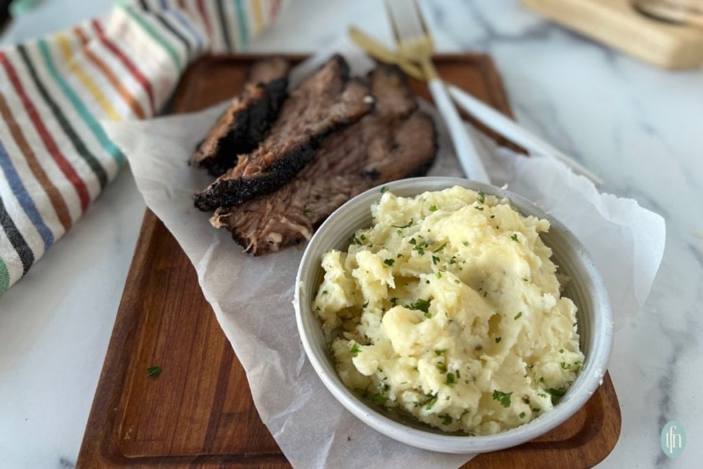 a bowl of mashed potatoes served alongside sliced smoked brisket on a wooden cutting board.
