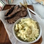 a bowl of mashed potatoes served alongside sliced smoked brisket on a wooden cutting board.