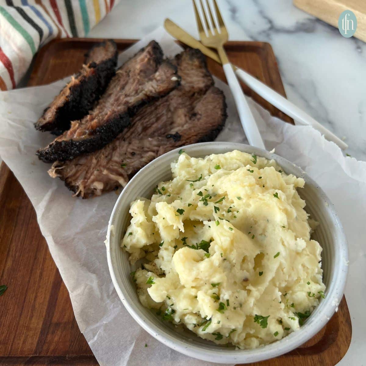 a bowl of mashed potatoes served alongside sliced smoked brisket on a wooden cutting board.
