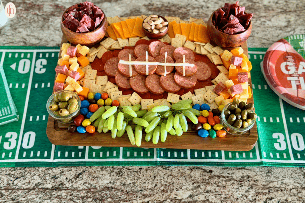 sliced meats arranged in the shape of a football and surrounded by grapes, olives, m and ms, almonds, mini saltine crackers, cubed meat and cheese, sliced cheese, mini pickles, and beef jerky.