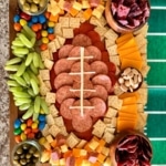 sliced meats arranged in the shape of a football and surrounded by grapes, olives, m and ms, almonds, mini saltine crackers, cubed meat and cheese, sliced cheese, mini pickles, and beef jerky.