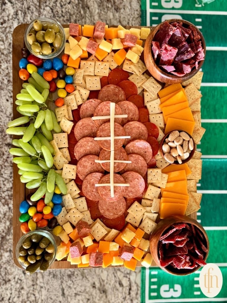 sliced meats arranged in the shape of a football and surrounded by grapes, olives, m and ms, almonds, mini saltine crackers, cubed meat and cheese, sliced cheese, mini pickles, and beef jerky.